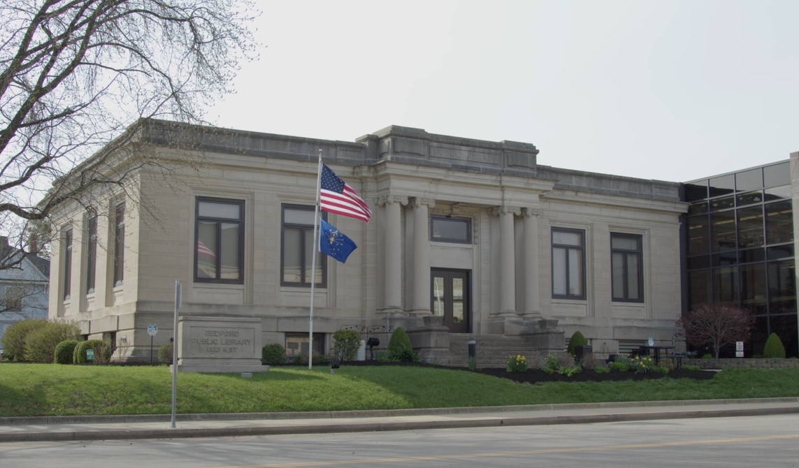 Square limestone building in the style of a Carnegie library, with four columns surrounding a stone step, landscaped flower beds top the sloping lawn. A United States and an Indiana state flag wave atop a flagpole next to a limestone Bedford Public Library sign.