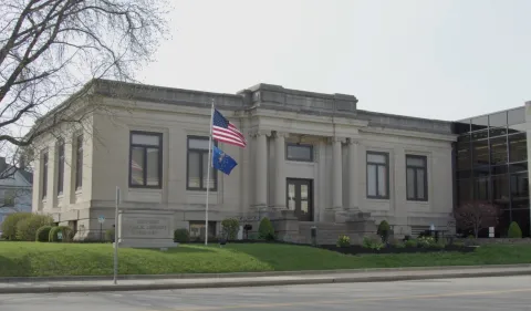 Square limestone building in the style of a Carnegie library, with four columns surrounding a stone step, landscaped flower beds top the sloping lawn. A United States and an Indiana state flag wave atop a flagpole next to a limestone Bedford Public Library sign.
